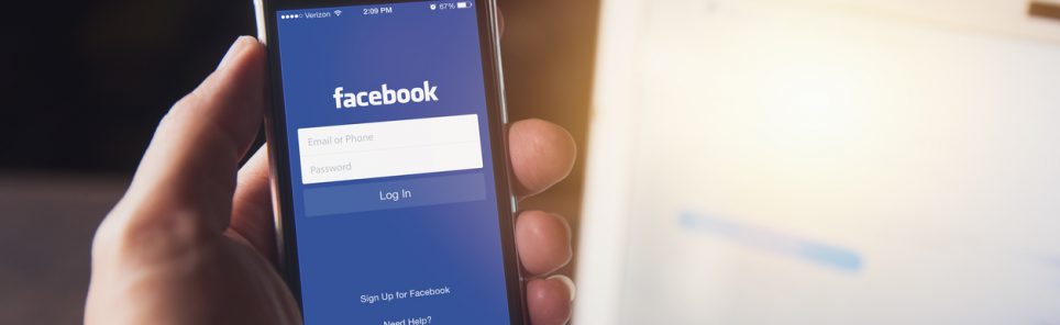 Facebook App on iPhone with computer laptop background closeup male hand hold social network on smart device concept.