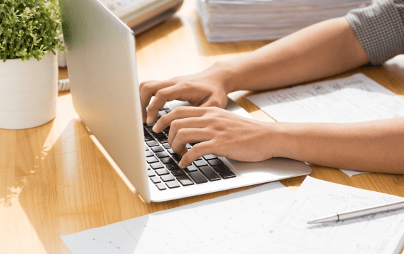 Person with rolled-up sleeves typing on a laptop next to architectural drawings and a mechanical pencil on a wooden desk, with a small potted plant beside the computer.