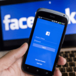 Person holding an HTC smartphone with the Facebook login screen, in front of a laptop displaying the Facebook logo.