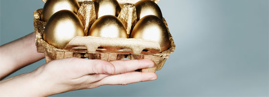 A hand holding a carton of golden eggs against a neutral grey background.