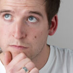 Young man with brown hair and blue eyes looking thoughtful, chin resting on hand, wearing a white t-shirt against a light gray background.