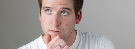 Young man with brown hair and blue eyes looking thoughtful, chin resting on hand, wearing a white t-shirt against a light gray background.