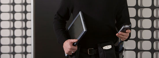 A person in business attire holding a laptop under one arm and a smartphone in the other hand, standing in front of a patterned wall.