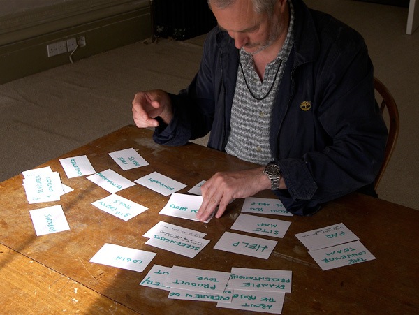 A man sitting at a wooden table organizing white cards with green handwriting on them.