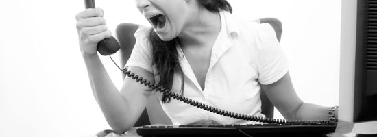 A woman in a white shirt shouting into a telephone receiver with a stretched cord, in front of a computer monitor, in a black and white image.