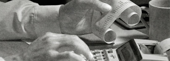 Hands of a person examining a printed receipt and operating a calculator, with coffee cups and papers on the desk, indicating a financial or accounting task.