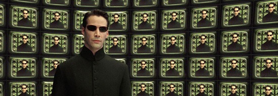 A man in sunglasses and a black outfit stands in front of a wall of television screens, each showing the same image of him.