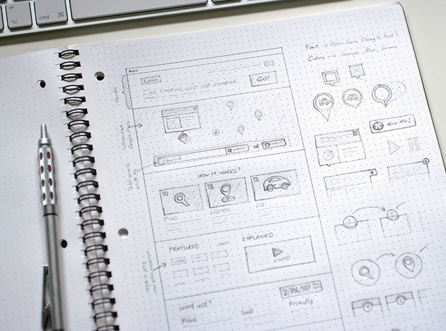 An open notebook with hand-drawn wireframe sketches for a website layout, annotations for font and color choices, a pen to the left, and a partial view of a keyboard at the top.