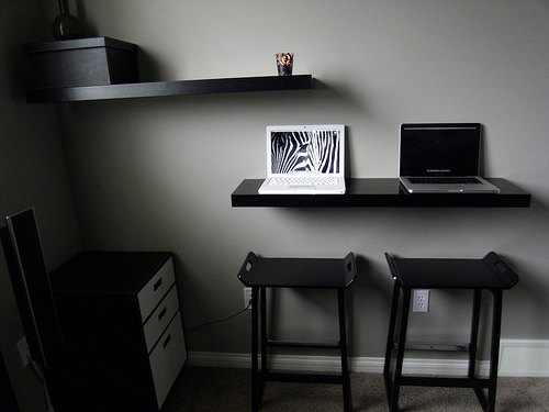 A modern home office setup with a black wall-mounted desk and floating shelf, a framed zebra print, a laptop, two black stools, and a three-drawer filing cabinet against a gray wall.