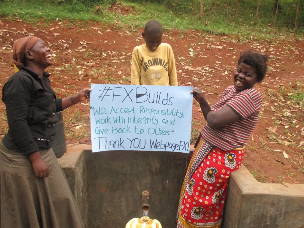 Two joyful women and a child standing by a water tap in a rural area, holding a sign with a message of gratitude and values, expressing thanks to a specific entity.