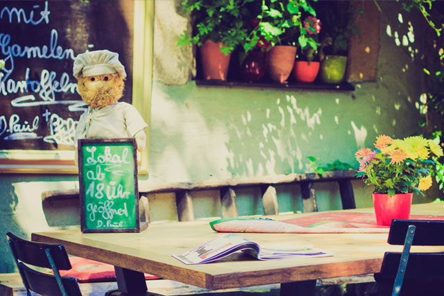 A cozy outdoor café setting with a wooden table featuring a chalkboard, a magazine, and a pot of flowers, with a chef figure standing behind the board and hanging plants on the wall.