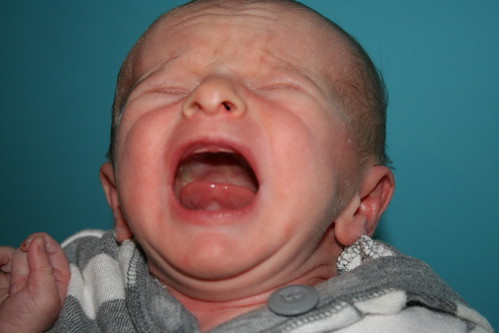 A newborn baby crying with eyes closed, wearing a grey and white striped garment against a blue background.