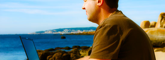 A person sitting by the seaside with a laptop, facing away from the camera and looking into the distance.