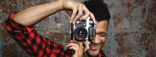 Smiling young man in a red and black checkered shirt taking a photo with a camera against a textured wall.