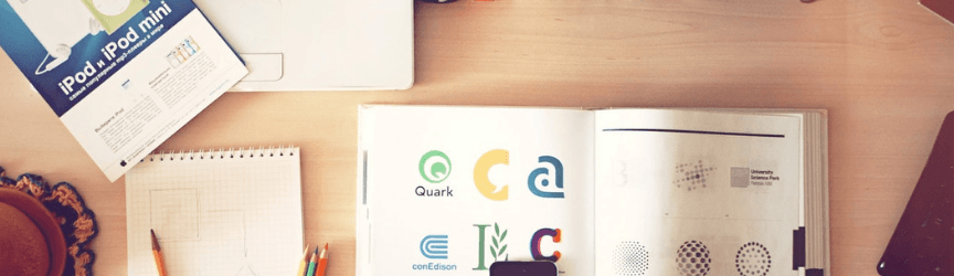 A desk with a spiral notebook, colored pencils, a box with 'iPod mini' text, an open magazine with colorful logos and 'E=mc²', and a partially visible laptop.