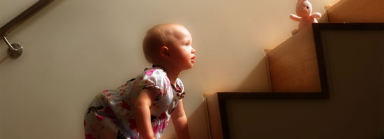 A young child on hands and knees gazes up at a plush toy sitting at the top of a staircase, illuminated by warm light.