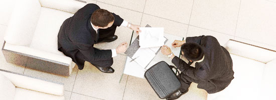 Two professionals in business attire sitting on a white sofa, reviewing documents and discussing, with a black briefcase beside them.