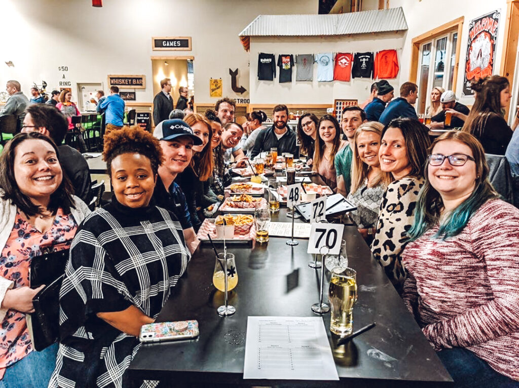 A group of people posing for a photo at a long table in a social setting with drinks and food, with signs for a whiskey bar and merchandise in the background.