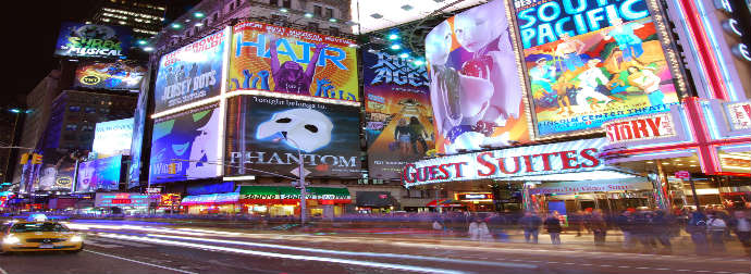 Night view of Times Square in New York City with illuminated billboards for Broadway shows, a yellow taxi in the foreground, and light streaks from moving traffic.