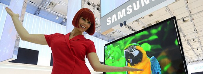 Humanoid robot with red hair presenting a Samsung flat-screen TV displaying a colorful parrot image.