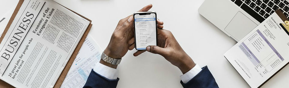 A person at a desk holding a smartphone with an email inbox displayed, surrounded by an open business newspaper, a laptop, and financial documents, suggesting a business or financial work setting.