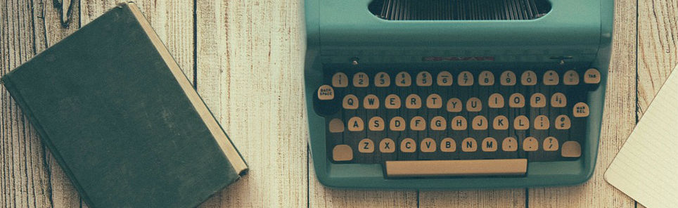 A vintage teal typewriter with a closed, worn hardcover book to its left and a blank piece of paper below it, all on a wooden surface.