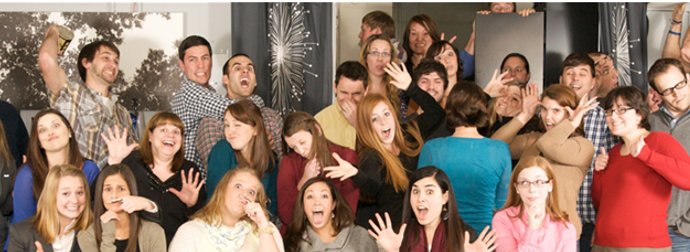 A group of young adults posing playfully for a group photo with some crouching in the front and others standing behind, making various gestures and expressions in an indoor setting.