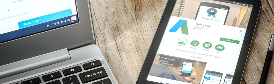 A laptop with Google search page and a tablet with Google AdWords page on Google Play Store, placed on a wooden desk with a stylus pen beside the tablet.