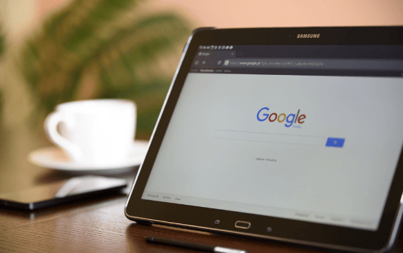 A Samsung tablet on a wooden table showing the Google search homepage, with a coffee cup and a blurred plant in the background.