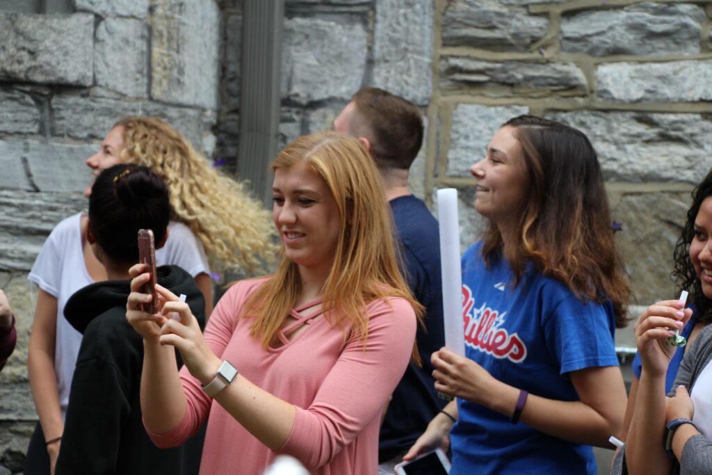 A young woman with blonde hair taking a selfie with her smartphone, surrounded by other smiling people at an outdoor social gathering.