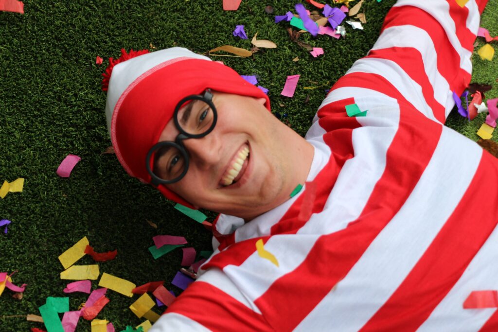 A person dressed as Waldo from 'Where's Waldo?', lying on grass with colorful confetti around, smiling at the camera.