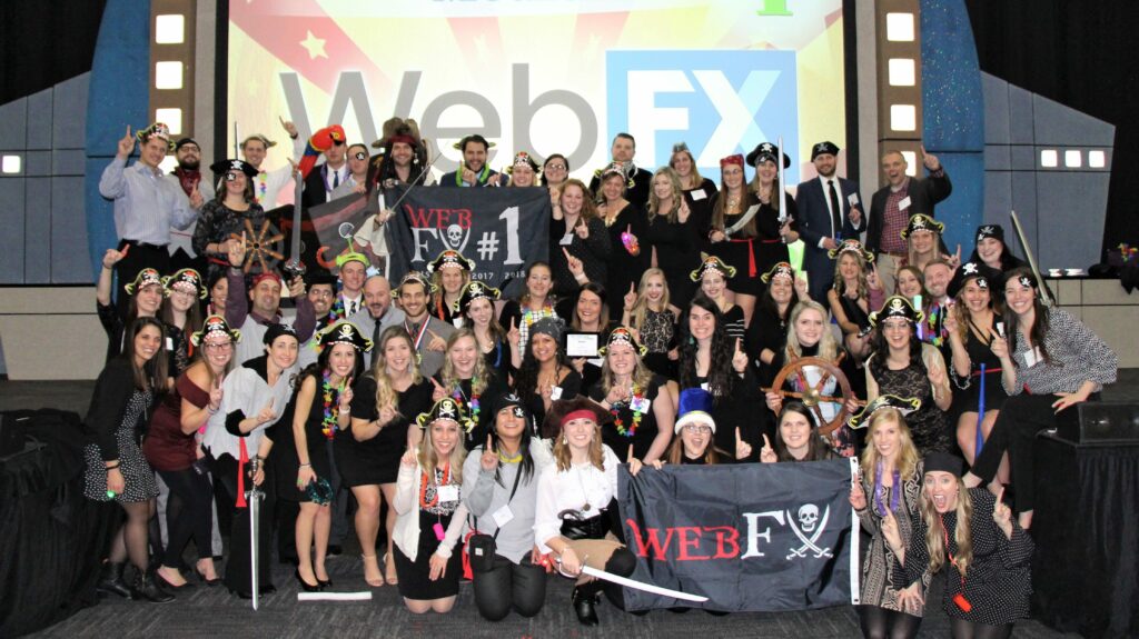 A group of people at a themed event, wearing pirate hats and holding props, celebrating in front of a 'Web FX' backdrop with a starry night graphic.