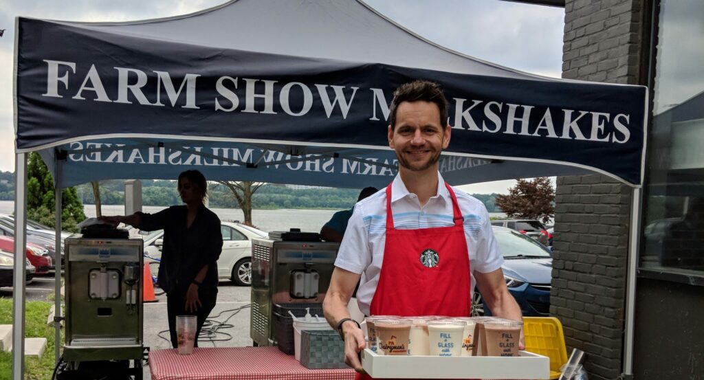 Man in a red Starbucks apron holding a carrier with four milkshakes at a 'FARM SHOW MILKSHAKES' stand, with milkshake machines and another worker in the background, at an outdoor event.