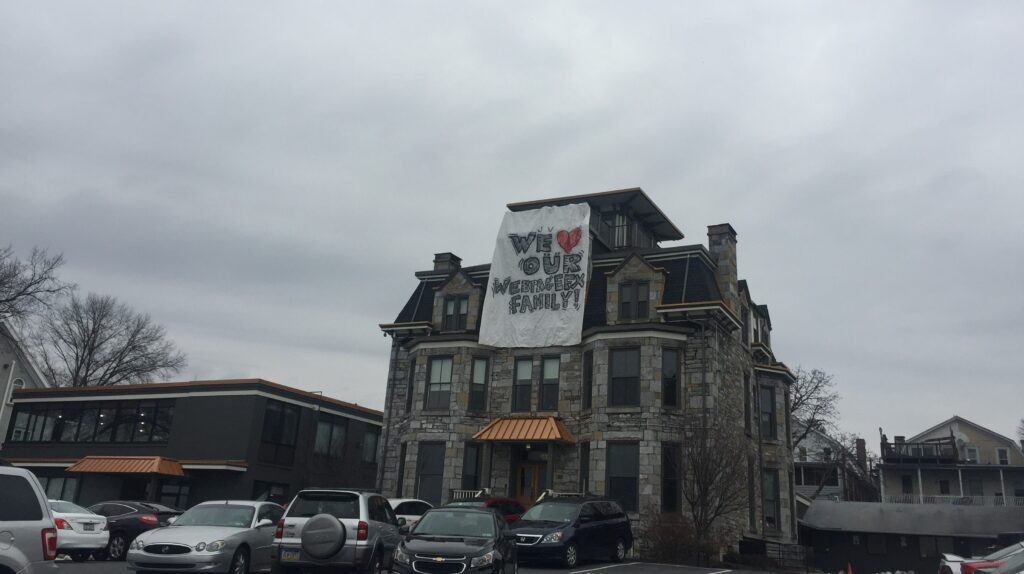 A large stone building with a banner reading 'WE ♥ OUR WEBERGEX FAMILY!' in front of an overcast sky with parked cars in the foreground.