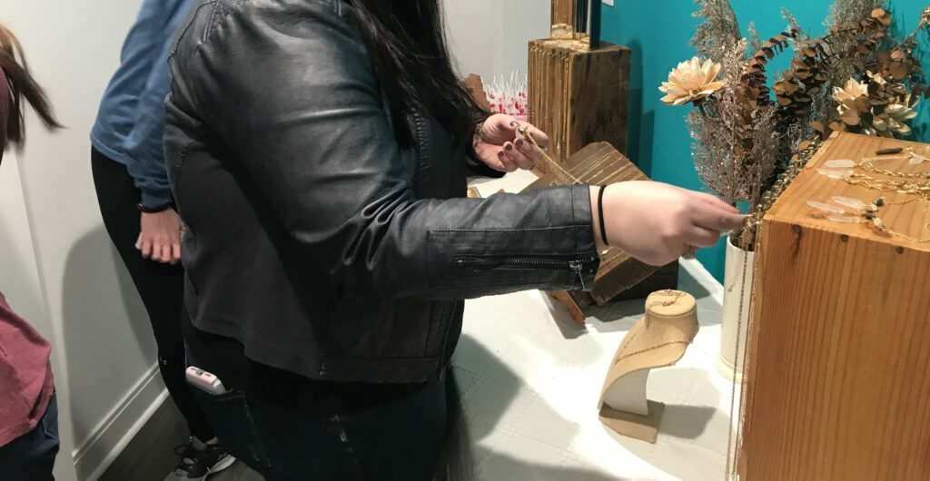 A person in a black leather jacket is browsing jewelry on a wooden display stand adorned with dried flowers in an indoor setting.