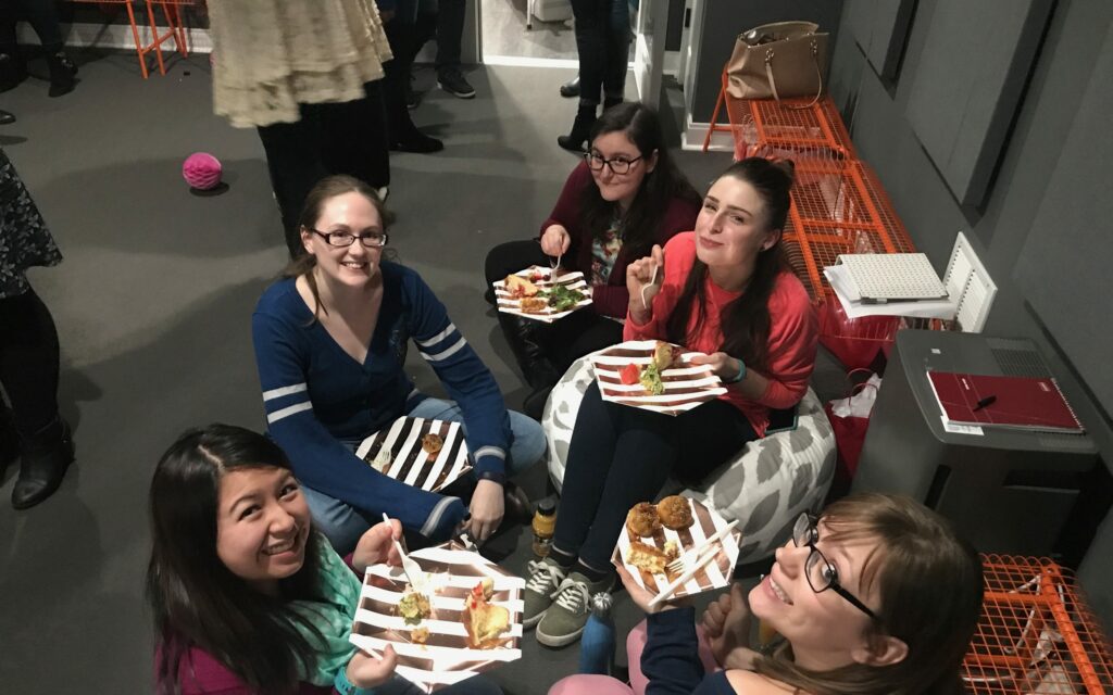 Five women sitting and enjoying food at an indoor gathering, surrounded by personal items and notebooks.