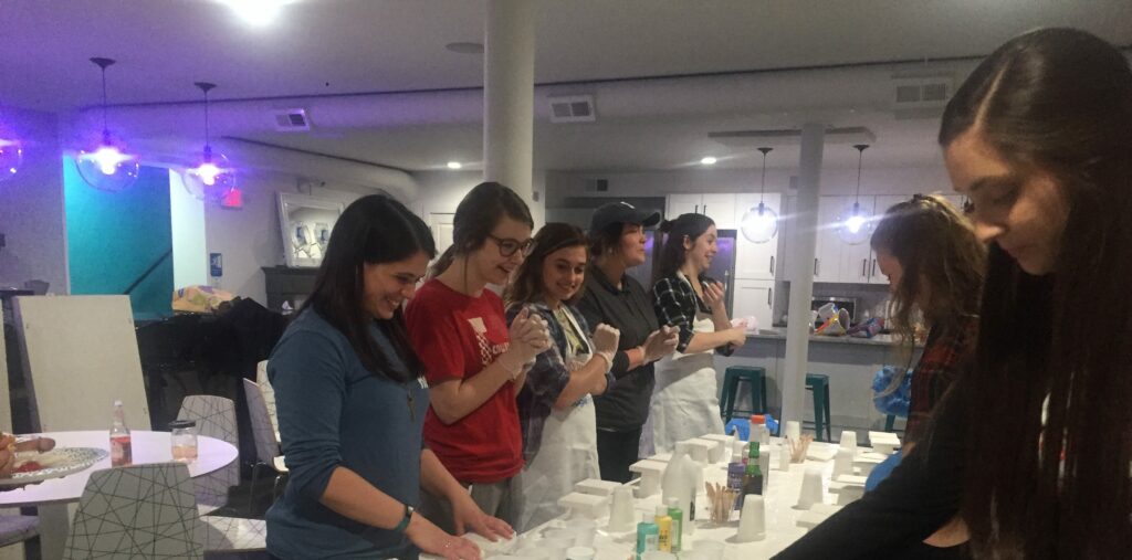 A group of people participating in a hands-on workshop, standing around a table with bottles and containers, some wearing aprons, in a well-lit indoor space with a kitchen in the background.