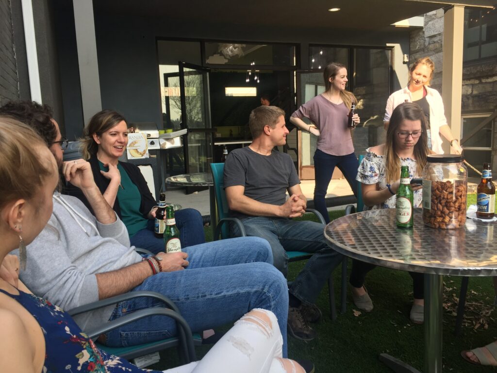 A group of people socializing on a patio with beers and snacks, including a woman smiling and holding a beer, a man listening attentively, and others chatting in the background.