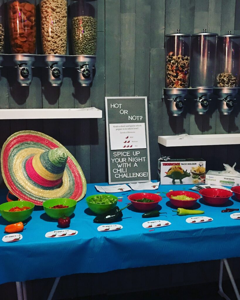 A table set up for a chili pepper challenge with labeled bowls of peppers, a sign reading 'Hot or Not?', snack dispensers above, a sombrero, a dinosaur-shaped taco holder, and a pair of gloves.