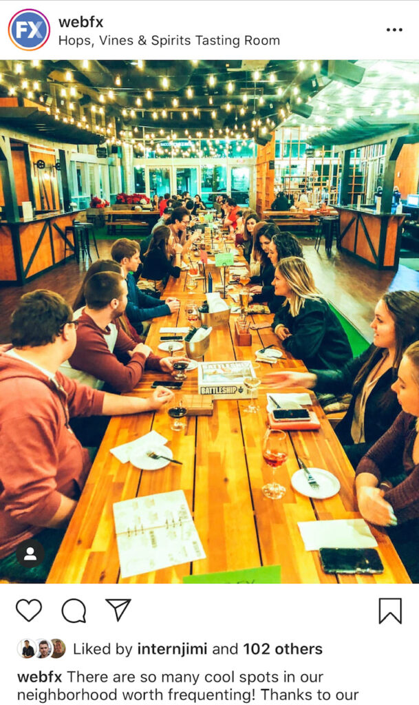 A group of people socializing at a long wooden table in a tasting room with string lights overhead, enjoying beverages and playing board games like Battleship, with a bar area in the background.