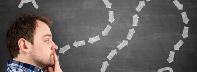 A man in a plaid shirt contemplating a series of chalk-drawn arrows curving above his head on a chalkboard, symbolizing a thought process or decision-making.