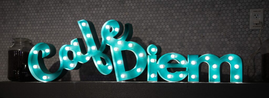 Illuminated turquoise 'carpe diem' cursive light-up sign on a shelf with a jar of coffee beans and a cup to the left, against a hexagonal tiled wall with an electrical outlet to the right.