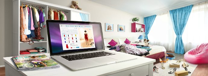 A laptop on a desk displaying a clothing website, with a bedroom in the background featuring a bed with colorful pillows, a closet, and blue curtains.