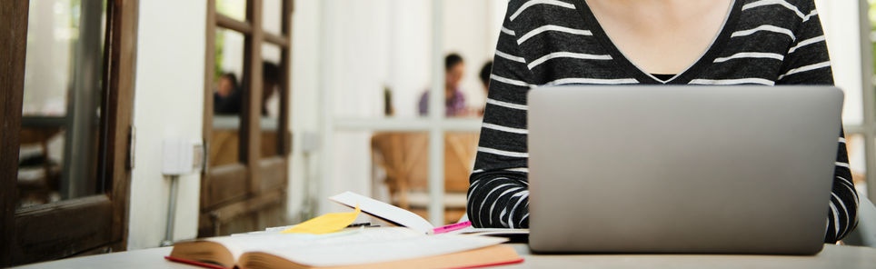 Person in a striped top working on a laptop with an open book and papers on the table in a well-lit room with blurred figures in the background.