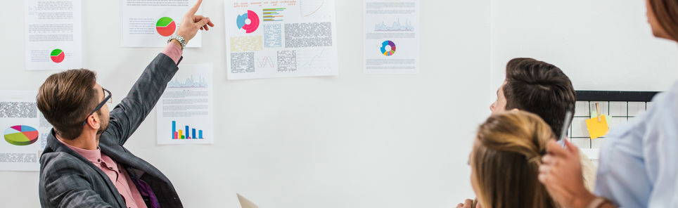 A man pointing at a pie chart during a presentation with colleagues looking on, surrounded by various charts and documents on a wall.