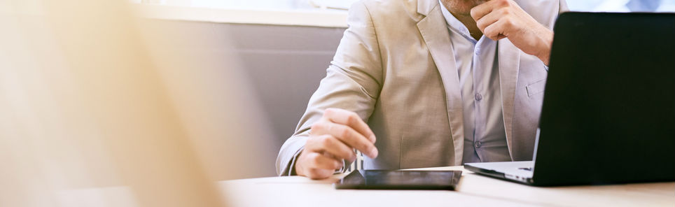 A man in a light suit jacket sitting at a desk with his chin resting on his hand, in front of a smartphone and an open laptop, appearing contemplative.