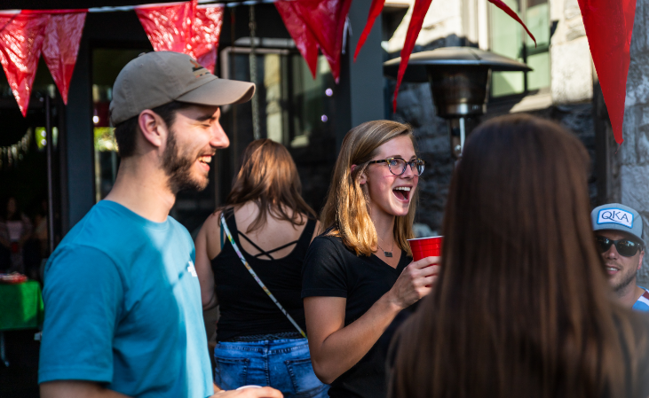 A group of people enjoying an outdoor social event, with a laughing woman holding a red cup in the foreground and a smiling man in a baseball cap to her left, under red triangular flags.