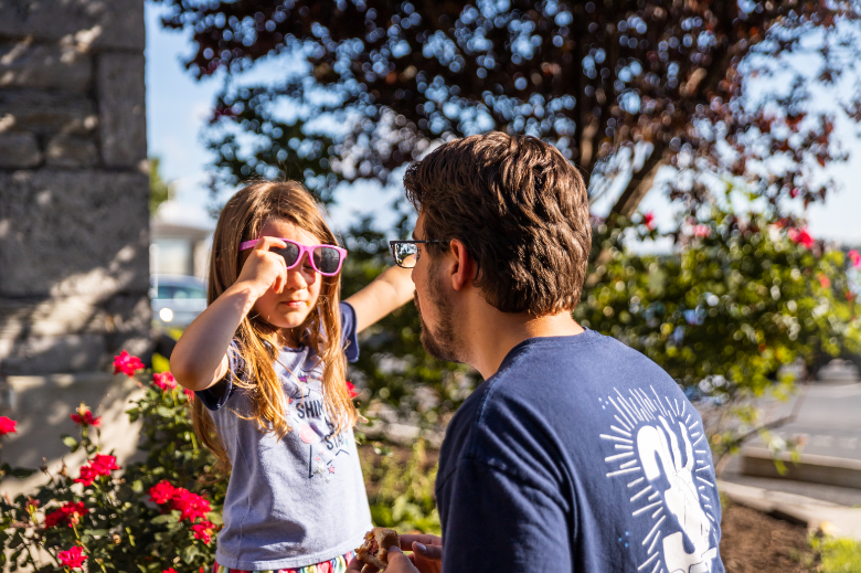 A young girl adjusting her pink sunglasses while looking at a bearded man in a blue t-shirt, both engaging in a conversation outdoors with red flowers and greenery in the background on a sunny day.