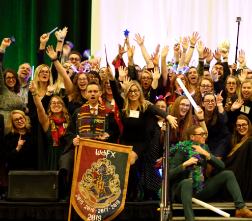 A lively group of people at an event, raising their hands with excitement, one holding a 'WebFX' banner, with a stage and green curtain in the background.