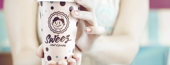 Close-up of a person's hands holding a paper cup with 'SweeZ sweet & orange' logo and polka dot design, with a blurred turquoise and pink background.